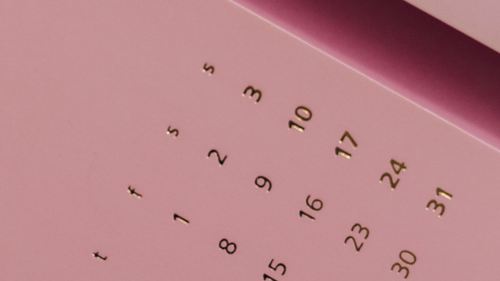 A dusty pink paper calendar photographed close up at an angle, its dates printed in gold and black