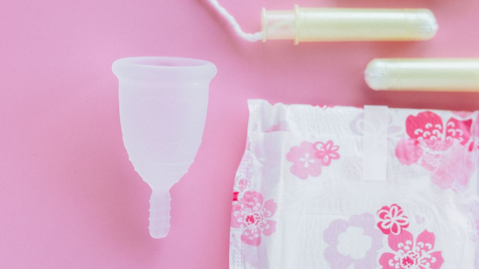 A menstrual cup, three tampons, and two wrapped pads laid out on a soft pink background