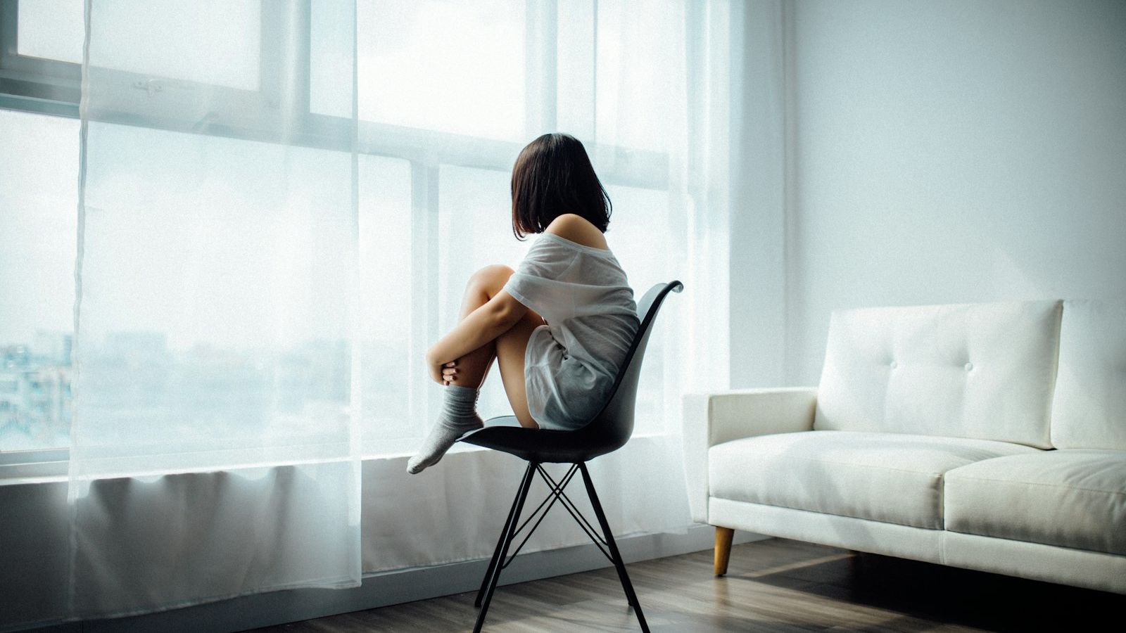 A woman sitting curled up on a chair by a bright, sheer-curtained window in a calm, light-filled room