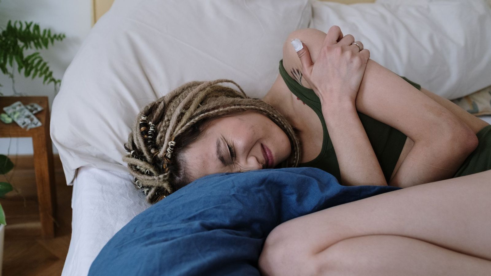 A young woman curled up on her side in bed first thing in the morning, eyes closed and face tensed with pain, holding her arms against her body