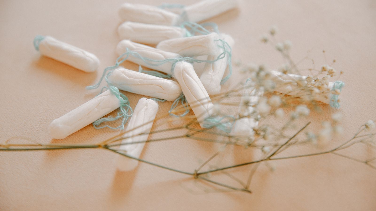 A scattered pile of cotton tampons with pale blue strings on a warm cream surface, beside a sprig of dried baby’s breath