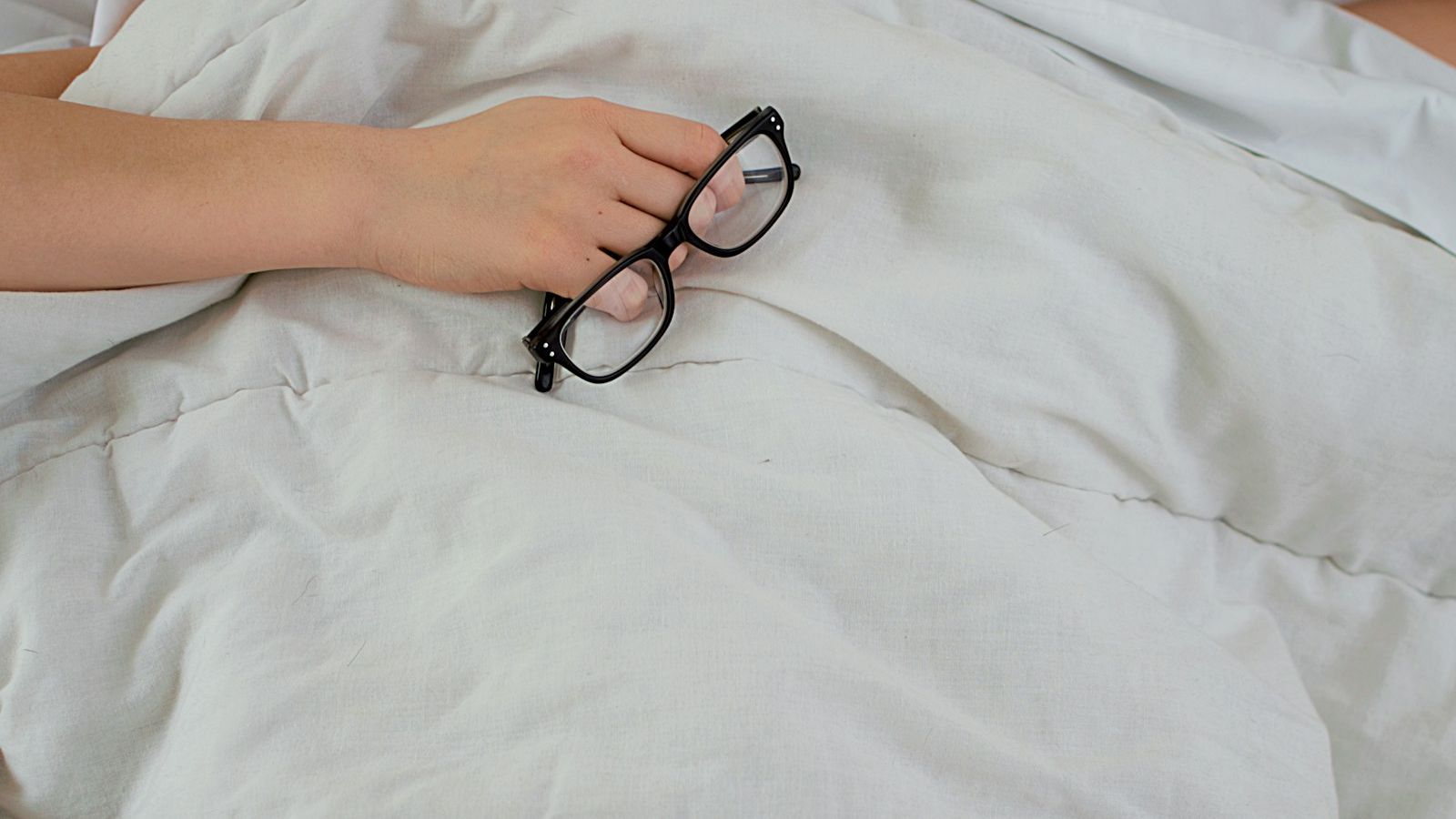 A person lying in bed with a pillow pulled over their face, holding a pair of black eyeglasses in one hand